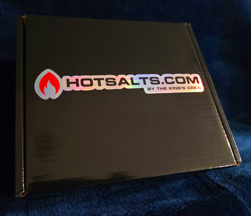 A black cardboard box with a holographic Hotsalts.com logo sticker on the front, resting on a dark blue textured fabric background.