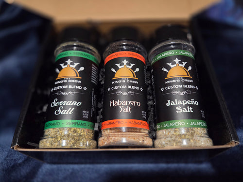 Three spice salt bottles from King's Crew Custom Blend arranged in a black gift box. The bottles are labeled Serrano Salt, Habanero Salt, and Jalapeño Salt, with green, orange, and white labels respectively, set against a dark blue fabric background.